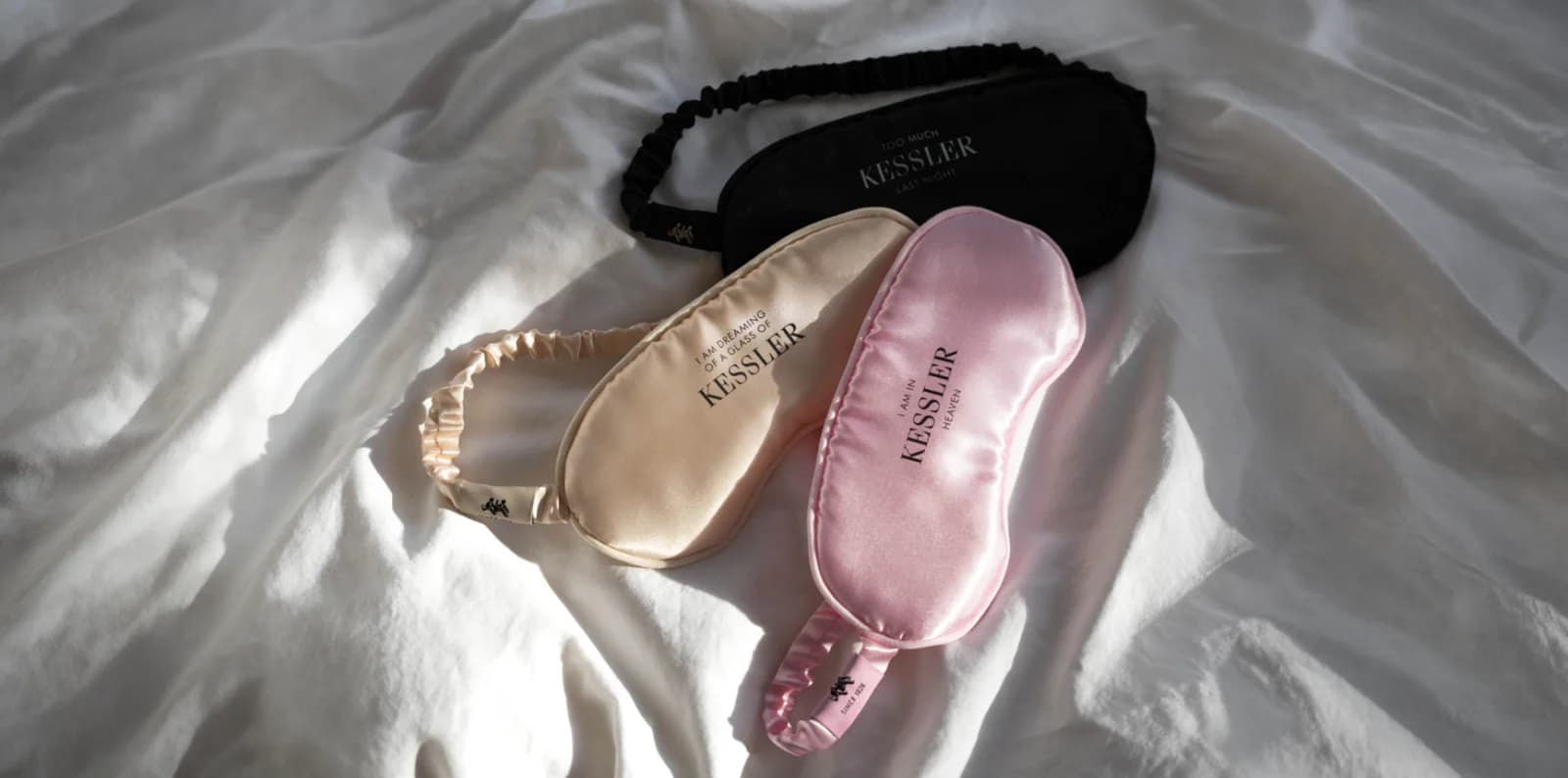 Drei Kessler-Schlafmasken aus Satin in Schwarz, Champagner und Rosé auf weißem Bettzeug