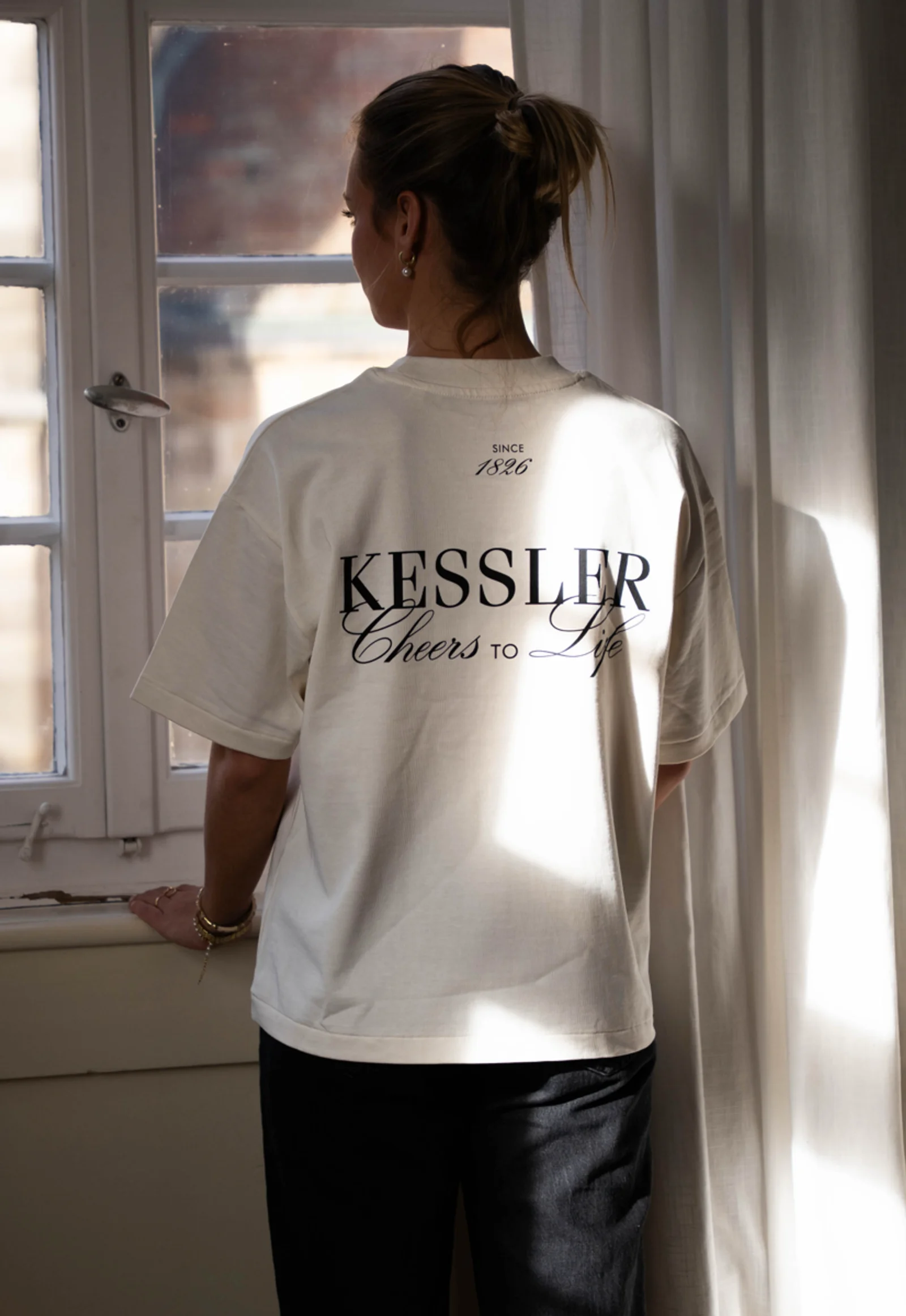 Cremeweißes Kessler-T-Shirt mit »Cheers to Life«-Rückenprint, getragen am Fenster